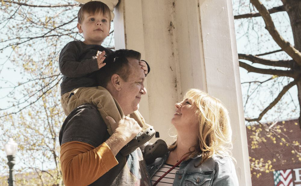 Two parents stand looking at each other while their small child rests on one of the parent's shoulders.